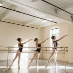 three dancers practicing on a portable ballet barre in a lit studio